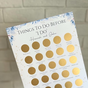 May include: A wedding planning checklist with gold scratch-off circles. The text reads "Things to do before 'I do'" and includes tasks like "Book Venue" and "Final Dress Fitting". Floral accents in blue and gold decorate the edges.