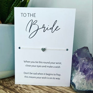 May include: White card with black text that reads "TO THE Bride". A white string bracelet with a silver heart charm is attached to the card. The text on the card reads "When you tie this round your wrist, close your eyes and make a wish. Don't be sad when it begins to fray, this means your wish is on its way."