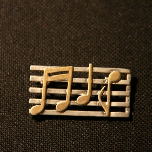 Könnte beinhalten: Eine silberne und goldene Musiknotennadel mit fünf Linien, die einen Notensystem darstellen. Die Noten sind stilisiert und haben einen Vintage-Look.