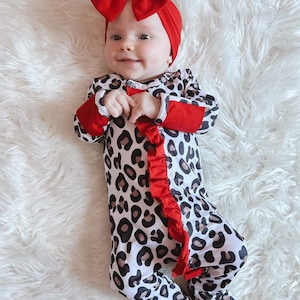 Puede incluir: Un bebé con un pijama de leopardo blanco y negro con ribetes rojos y una diadema roja con lazo.