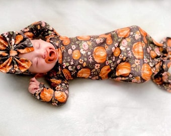 Può includere: Un neonato con un completo a tema zucca, che include una fascia con fiocco abbinata e un vestito annodato. Il completo presenta uno sfondo marrone scuro con zucche arancioni e dettagli floreali. Il bambino indossa anche un ciuccio.