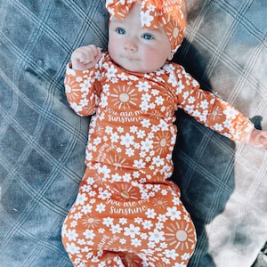 Puede incluir: Un bebé con un mono de manga larga de color naranja y blanco con estampado floral con el texto "You are my sunshine" repetido en todo el estampado. El bebé lleva una diadema a juego de color naranja y blanco con estampado floral y un gran lazo.