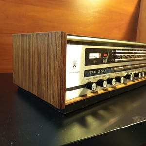 Könnte beinhalten: Ein Vintage RTV 350 Stereo-Receiver mit einer silberfarbenen Frontplatte, mehreren schwarzen Drehreglern, einer Radiofrequenzanzeige und einem Gehäuse mit Holzmaserung an der Seite. Das Gerät enthält ein kleines Logo und den Hinweis Made in Germany, mit einem schwarzen Sockel und horizontalem Design.