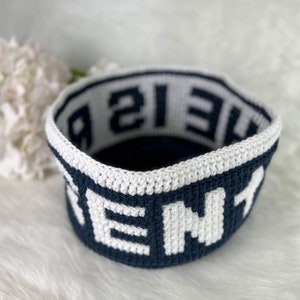 Op de afbeelding: Een gehaakte mand met een donkerblauwe basis en een witte rand. Het woord "HOME" is in wit op de donkerblauwe basis geschreven. Het woord "THE" is in donkerblauw op de witte rand geschreven.