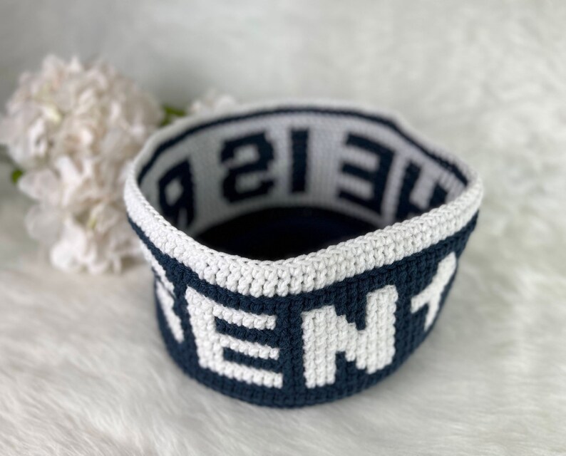 Op de afbeelding: Een gehaakte mand met een donkerblauwe basis en een witte rand. Het woord "HOME" is in wit op de donkerblauwe basis geschreven. Het woord "THE" is in donkerblauw op de witte rand geschreven.