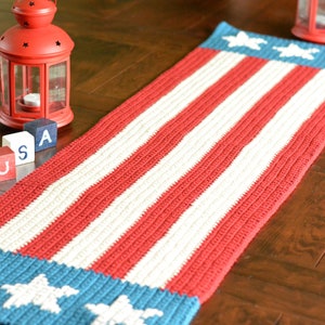 May include: A red, white, and blue crocheted table runner with stars on the ends. The runner is on a wooden table with red, white, and blue blocks that spell "USA".