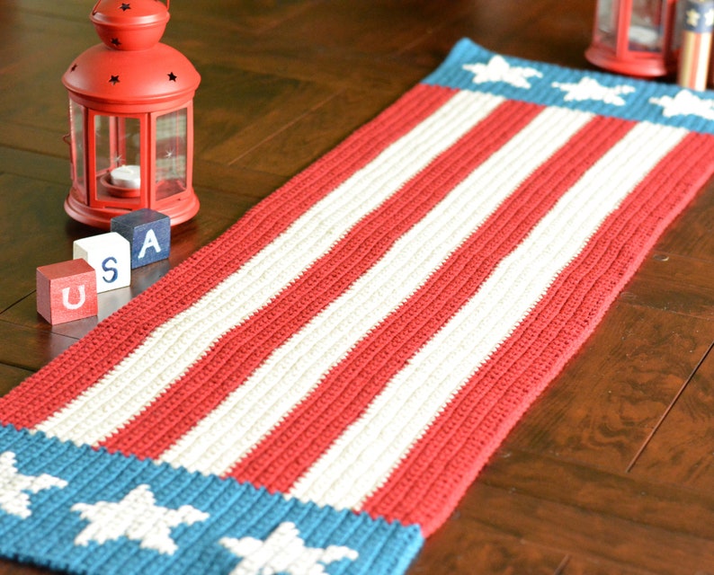 May include: A red, white, and blue crocheted table runner with stars on the ends. The runner is on a wooden table with red, white, and blue blocks that spell "USA".