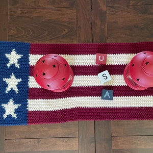 May include: A red, white, and blue crocheted table runner with three wooden blocks spelling "USA". Two red lanterns with black star cutouts are placed on the runner.