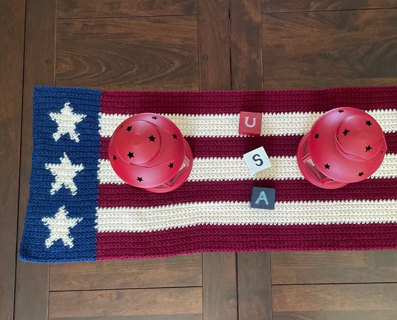 May include: A red, white, and blue crocheted table runner with three wooden blocks spelling "USA". Two red lanterns with black star cutouts are placed on the runner.