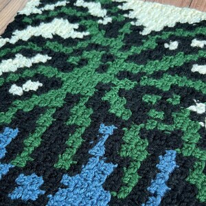 May include: A close-up of a crocheted square with a geometric pattern in shades of green, blue, black, and white.