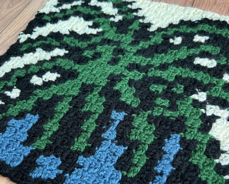 May include: A close-up of a crocheted square with a geometric pattern in shades of green, blue, black, and white.