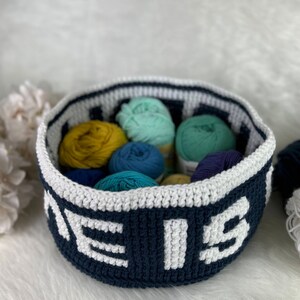 Op de afbeelding: Een donkerblauwe gehaakte mand met witte letters die "The" en "is" op de voorkant lezen. De mand is gevuld met kleurrijke garenbollen in tinten blauw, groen en geel.