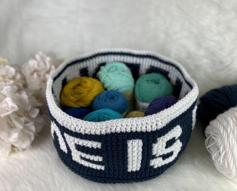 Op de afbeelding: Een donkerblauwe gehaakte mand met witte letters die "The" en "is" op de voorkant lezen. De mand is gevuld met kleurrijke garenbollen in tinten blauw, groen en geel.