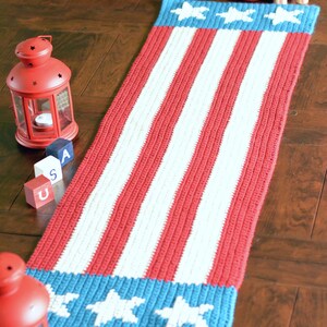 May include: A red, white, and blue crocheted table runner with stars on the ends. The runner is on a wooden floor with red lanterns and wooden blocks that spell "USA".