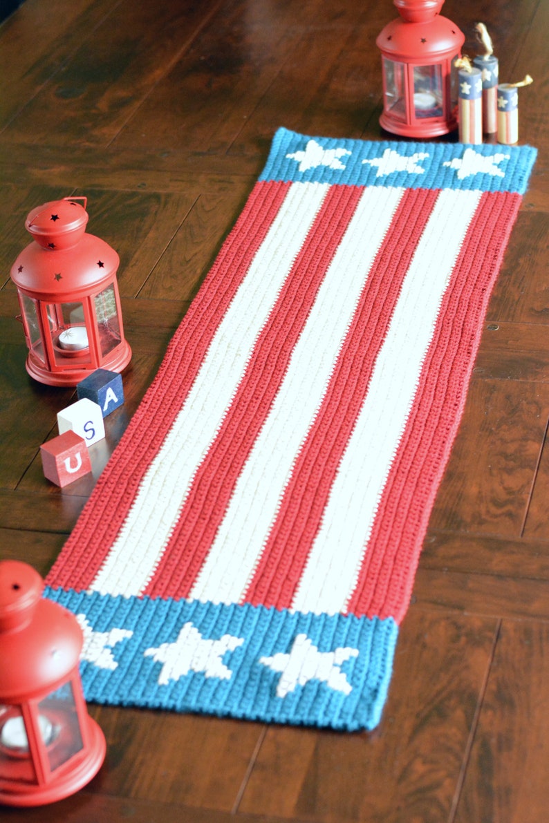 May include: A red, white, and blue crocheted table runner with stars on the ends. The runner is on a wooden floor with red lanterns and wooden blocks that spell "USA".