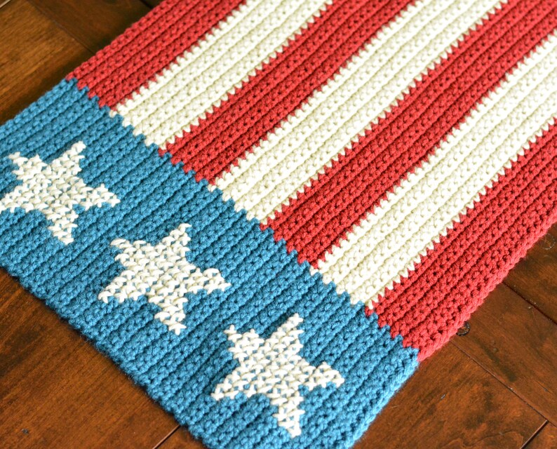 May include: A crocheted table runner in red, white, and blue with a design resembling the American flag. The runner features white stars on a blue background and red and white stripes.