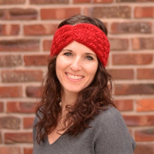 Op de afbeelding: Een vrouw met lang, krullend bruin haar draagt een rood gebreid hoofdband. Het hoofdband is een gedraaid ontwerp met een textuurpatroon. De vrouw glimlacht en kijkt naar de camera.