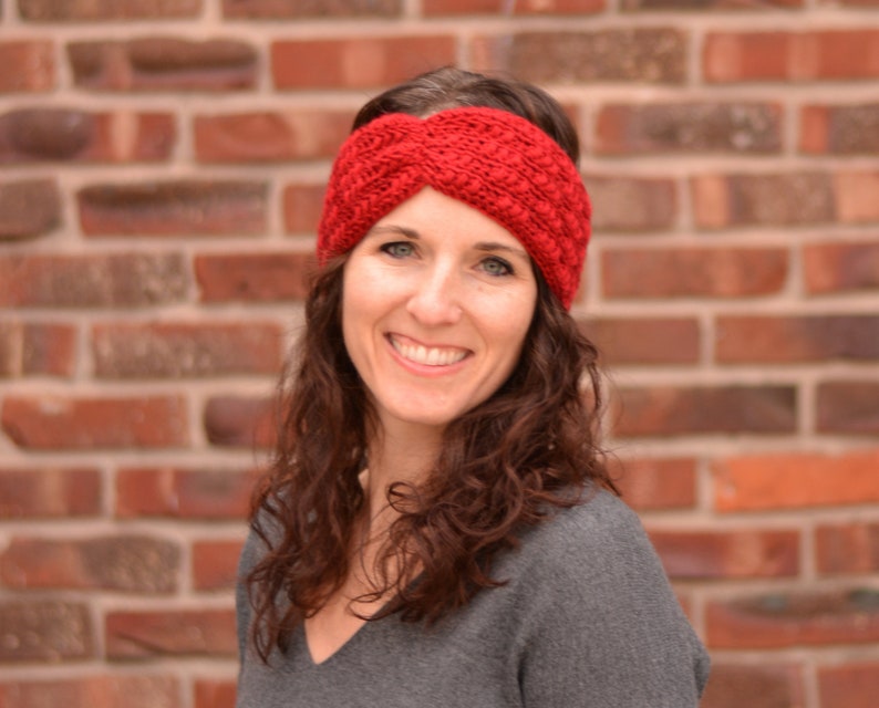 Op de afbeelding: Een vrouw met lang, krullend bruin haar draagt een rood gebreid hoofdband. Het hoofdband is een gedraaid ontwerp met een textuurpatroon. De vrouw glimlacht en kijkt naar de camera.
