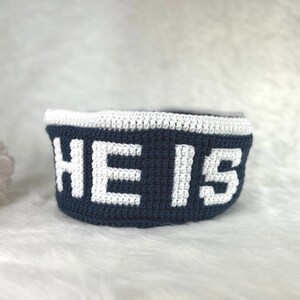 Op de afbeelding: Een blauwe gehaakte mand met een witte gehaakte rand en het woord "HE IS" in wit.