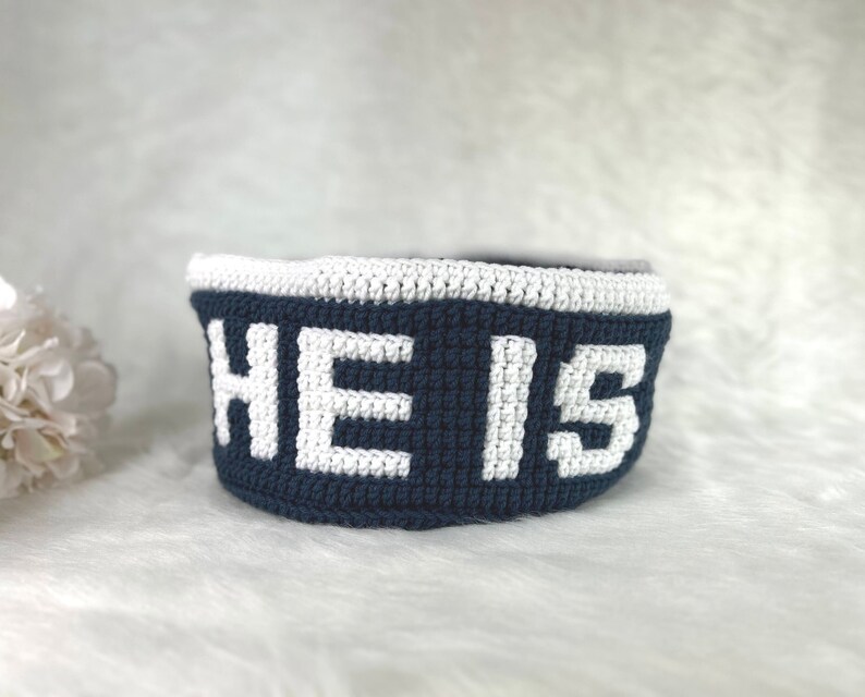 Op de afbeelding: Een blauwe gehaakte mand met een witte gehaakte rand en het woord "HE IS" in wit.