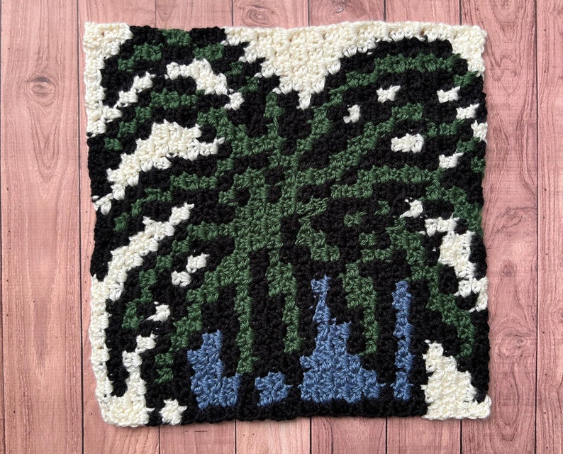 May include: A square crochet pattern with a green, black, and white design. The pattern is made with different colored yarn and has a geometric design.