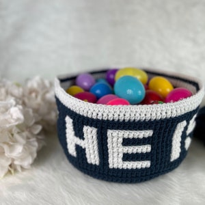 Op de afbeelding: Een blauwe gehaakte mand met witte letters die "HIER" spellen, is gevuld met kleurrijke plastic paaseieren.