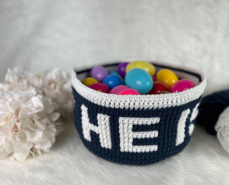 Op de afbeelding: Een blauwe gehaakte mand met witte letters die "HIER" spellen, is gevuld met kleurrijke plastic paaseieren.