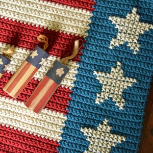 May include: A crocheted American flag rug with red, white, and blue stripes. The rug has three small wooden stars and stripes ornaments on top of it.