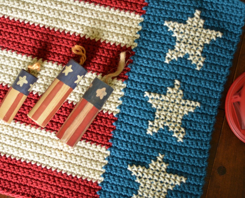 May include: A crocheted American flag rug with red, white, and blue stripes. The rug has three small wooden stars and stripes ornaments on top of it.