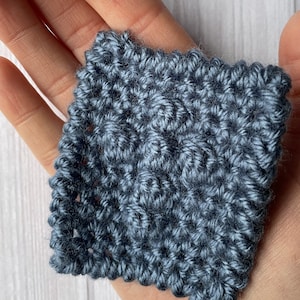 May include: A close-up of a small, blue crocheted square. The square is made with a textured stitch and has a slightly fuzzy appearance.
