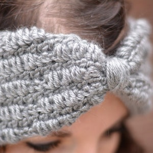 Op de afbeelding: Een grijs gehaakt hoofdband met een strikdetail. Het hoofdband wordt gedragen door een persoon met bruin haar.