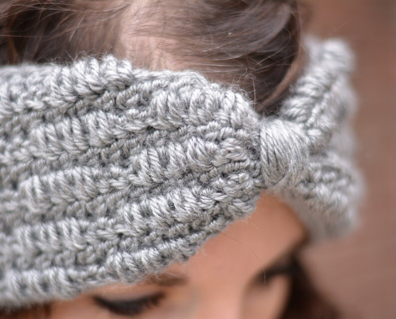 Op de afbeelding: Een grijs gehaakt hoofdband met een strikdetail. Het hoofdband wordt gedragen door een persoon met bruin haar.