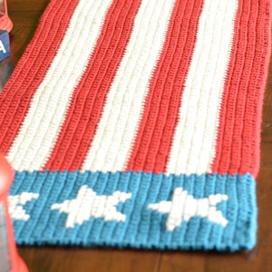 May include: A crocheted table runner in red, white, and blue with a star design. The runner is made with a textured stitch and has a patriotic theme.