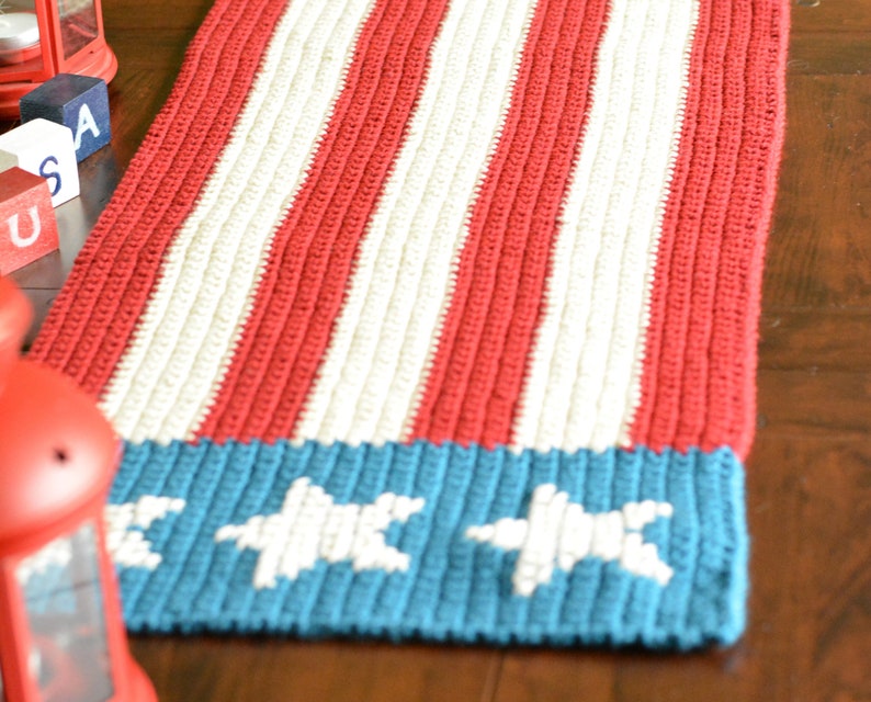 May include: A crocheted table runner in red, white, and blue with a star design. The runner is made with a textured stitch and has a patriotic theme.