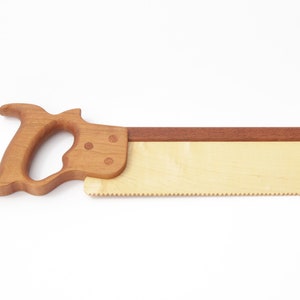 Peut inclure: Une scie à main en bois jouet avec une poignée brune et une lame brun clair. La lame a une bande rouge le long du dessus.