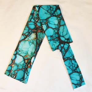 May include: A turquoise-colored, rectangular piece of material with a marbled pattern. The material has a network of dark brown veins and gold accents. The piece is folded in an angled shape, with a smaller, detached section extending from the main body.