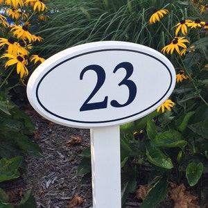 May include: White oval sign with black numbers "23" on a white wooden post. The sign is in a garden setting with yellow flowers.