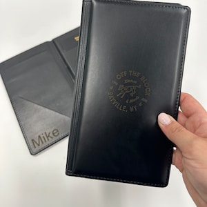 May include: Black leather menu holder with the text "OFF THE BLOCK Kitchen & Meats" and a cow graphic. The holder is embossed with the restaurant's name and location, Sayville, NY. The open holder has the name "Mike" engraved.