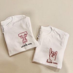 May include: Two white t-shirts with red embroidered designs. The first shirt has a red embroidered "T" with the words "wreck em" underneath. The second shirt has a red embroidered hand making the "hook 'em horns" gesture with the words "hook em" underneath.