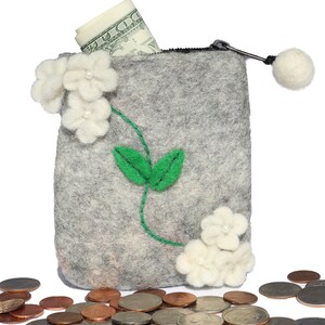 Puede incluir: Una cartera de monedas de fieltro gris con cierre de cremallera y un pomp&oacute;n blanco. La cartera est&aacute; decorada con flores de fieltro blanco y hojas de fieltro verde. Un billete de d&oacute;lar enrollado sobresale de la parte superior de la cartera. Hay varias monedas esparcidas alrededor de la cartera.