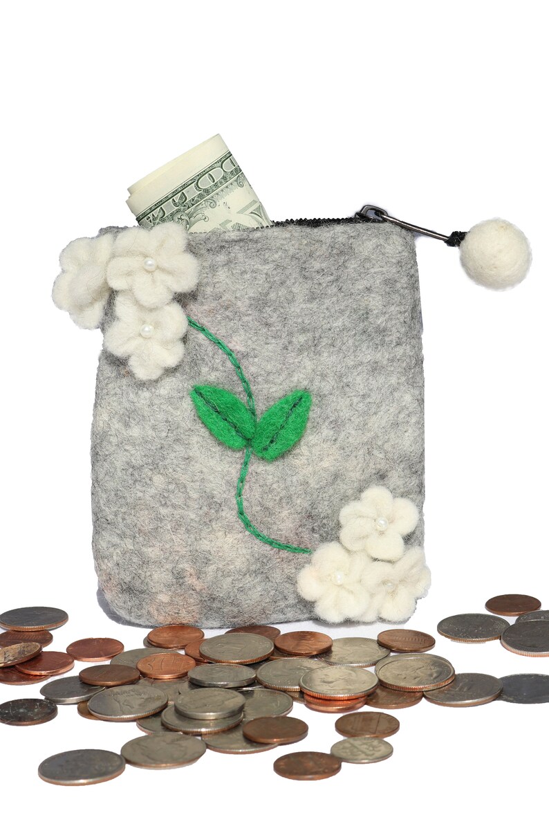 Puede incluir: Una cartera de monedas de fieltro gris con cierre de cremallera y un pomp&oacute;n blanco. La cartera est&aacute; decorada con flores de fieltro blanco y hojas de fieltro verde. Un billete de d&oacute;lar enrollado sobresale de la parte superior de la cartera. Hay varias monedas esparcidas alrededor de la cartera.