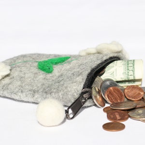 Puede incluir: Una bolsa de fieltro gris con un dise&ntilde;o de flor blanca y hoja verde. La bolsa est&aacute; abierta y se derraman billetes de d&oacute;lar estadounidense y monedas estadounidenses.