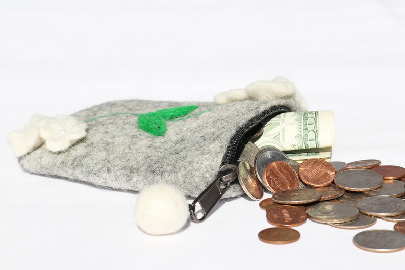 Puede incluir: Una bolsa de fieltro gris con un dise&ntilde;o de flor blanca y hoja verde. La bolsa est&aacute; abierta y se derraman billetes de d&oacute;lar estadounidense y monedas estadounidenses.