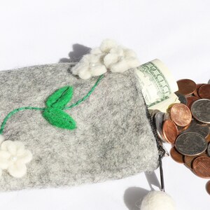 Puede incluir: Una cartera de monedas de fieltro gris con flores de fieltro blanco y hojas de fieltro verde. La cartera est&aacute; abierta y se caen monedas. La cartera tiene una cremallera y un borla de fieltro blanco.