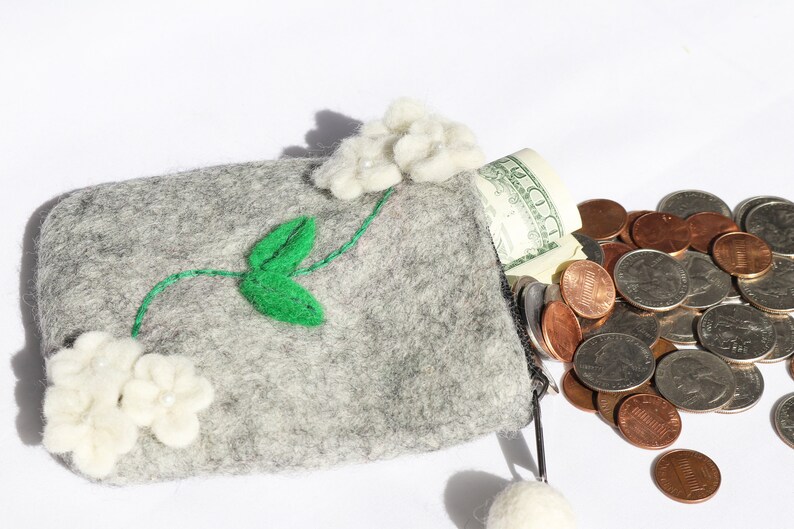 Puede incluir: Una cartera de monedas de fieltro gris con flores de fieltro blanco y hojas de fieltro verde. La cartera est&aacute; abierta y se caen monedas. La cartera tiene una cremallera y un borla de fieltro blanco.