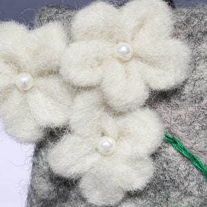 Puede incluir: Tres flores de fieltro blanco con centros de perlas est&aacute;n unidas a un fondo de fieltro gris. Un hilo verde est&aacute; cosido a lo largo de la parte inferior de las flores.