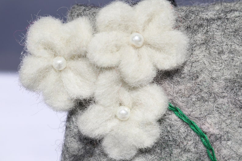 Puede incluir: Tres flores de fieltro blanco con centros de perlas est&aacute;n unidas a un fondo de fieltro gris. Un hilo verde est&aacute; cosido a lo largo de la parte inferior de las flores.