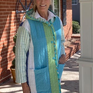 May include: A quilted coat with a hood, featuring a patchwork design of blue, green, and white vertical stripes and squares. The coat has two pockets and is buttoned up the front. The coat is worn over a white collared shirt.