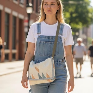 May include: A light blue denim overall with a white t-shirt and a cream-colored shoulder bag. The bag features a geometric pattern in shades of peach, blue, and cream. The bag has a green strap. The person is walking down a city street.