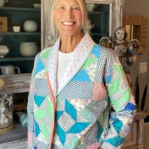 May include: A quilted jacket featuring a patchwork design of various fabric patterns in shades of blue, green, pink, and white. The garment has a shawl collar with a pink polka dot lining, long sleeves, and a straight hem, constructed from stitched fabric squares.